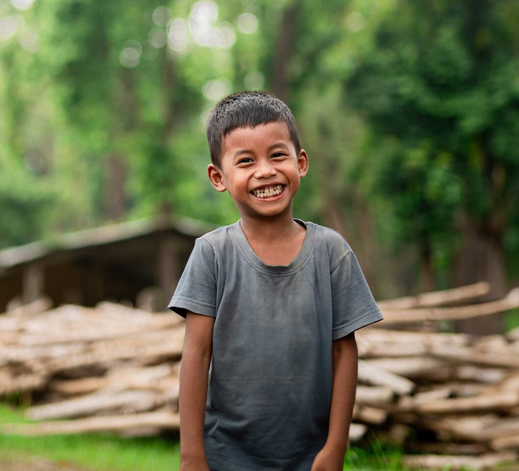 Boy standing with big smile in front of logs and greenery.